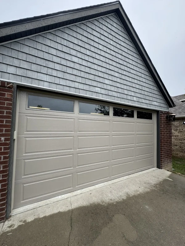 Elite Garage Door Services of Northwest ArkansasA beige residential garage door with windows in a brick house, showcasing a recent garage door installation in Northwest Arkansas, emphasizing local and professional service.