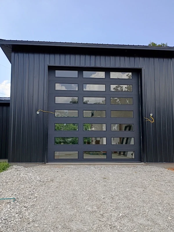 Elite Garage Door Services of Northwest ArkansasA sleek, black overhead garage door with glass panels is installed in a modern building, reflecting the surrounding trees and sky. The installation showcases professional garage door service in Northwest Arkansas.