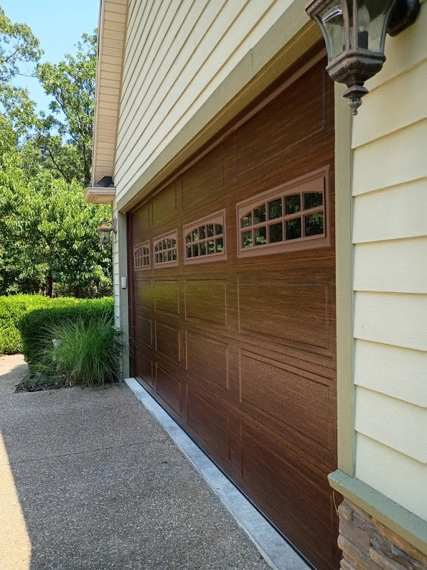 Elite Garage Door Services of Northwest ArkansasA newly installed residential garage door with wooden panels is seen in a home setting, surrounded by greenery. This professional garage door installation in Northwest Arkansas enhances curb appeal and functionality.