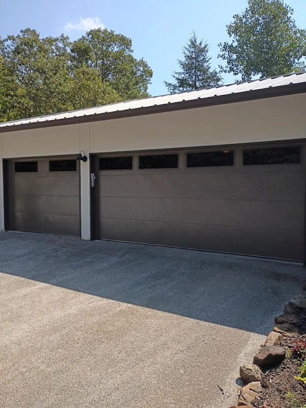 Elite Garage Door Services of Northwest ArkansasA beige house with two large brown overhead garage doors, surrounded by trees. These residential garage doors are part of a reliable garage door service in Northwest Arkansas, offering professional installation and repair to local homeowners.