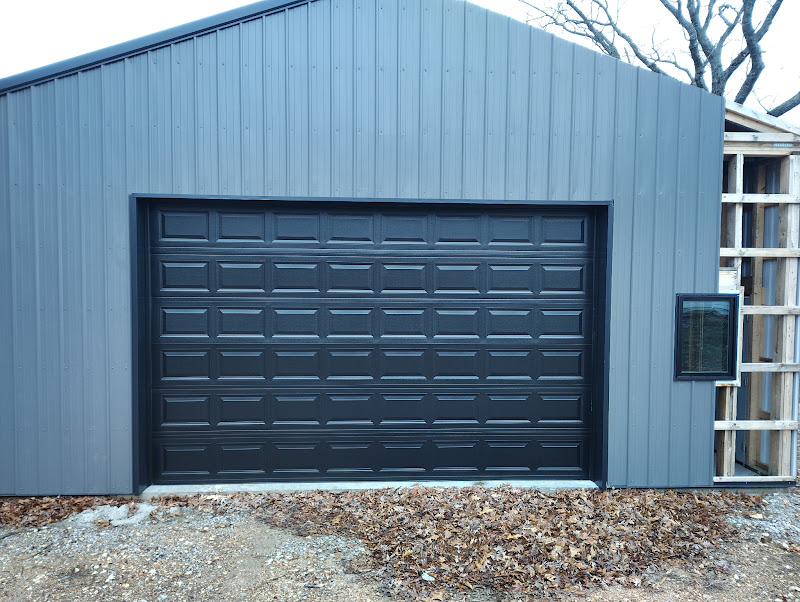 Elite Garage Door Services of Northwest ArkansasA newly installed black overhead garage door on a gray building, surrounded by autumn leaves, exemplifies the quality of garage door installation services in Northwest Arkansas, highlighting local expertise and reliability for residential needs.