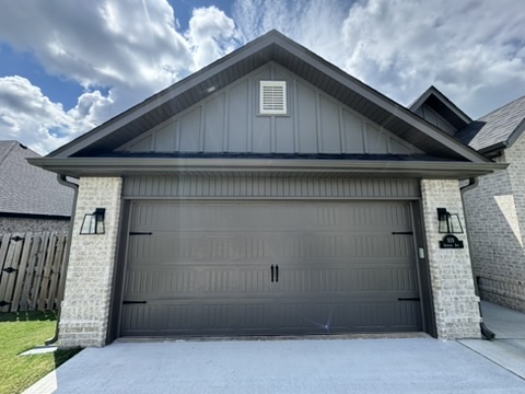 Elite Garage Door Services of Northwest ArkansasA modern, residential garage door in Northwest Arkansas is shown fully closed. The door is steel with a wood grain finish, flanked by brick walls under a clear blue sky. Ideal for reliable garage door service in NWA.