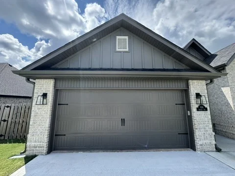 Elite Garage Door Services of Northwest ArkansasA modern, residential garage door in Northwest Arkansas is shown fully closed. The door is steel with a wood grain finish, flanked by brick walls under a clear blue sky. Ideal for reliable garage door service in NWA.