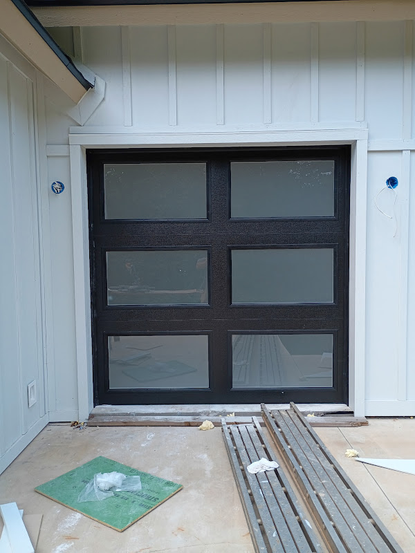 Elite Garage Door Services of Northwest ArkansasA newly installed black residential garage door with frosted glass panels is seen in a white exterior setting, showcasing professional garage door installation in Northwest Arkansas. Construction materials are visible on the ground.