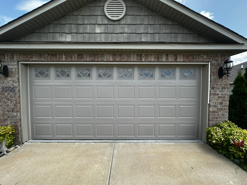 Elite Garage Door Services of Northwest ArkansasA beige residential garage door with decorative window panels, set in a brick house in Northwest Arkansas. The driveway is clean and well-maintained, reflecting the professional garage door service available in Rogers AR.