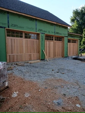 Elite Garage Door Services of Northwest ArkansasA newly constructed house in Northwest Arkansas features three wooden residential garage doors under installation, showcasing professional and reliable garage door service. The green house wrap is visible, and gravel covers the driveway area.