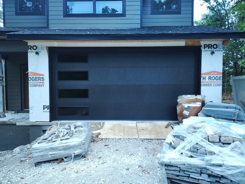 Elite Garage Door Services of Northwest ArkansasA newly installed black residential garage door is seen on a house under construction in Northwest Arkansas, surrounded by building materials. The door features four vertical windows, highlighting a professional garage door installation.