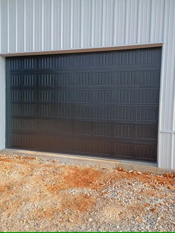 Elite Garage Door Services of Northwest ArkansasA newly installed black overhead garage door on a metal building in Northwest Arkansas. The area around the door shows gravel and red dirt, indicative of construction. This represents professional garage door service in the NWA region.