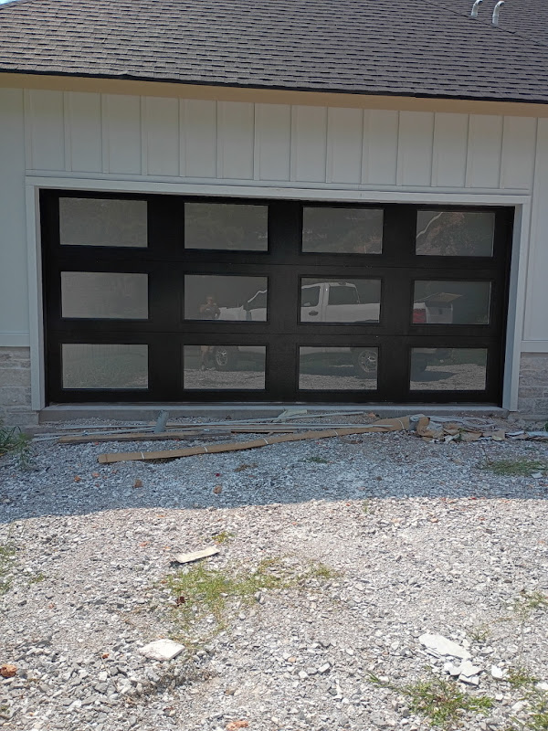 Elite Garage Door Services of Northwest ArkansasA newly installed black residential garage door with windows in a white house, surrounded by gravel and construction debris, showcasing professional garage door installation services in Northwest Arkansas.