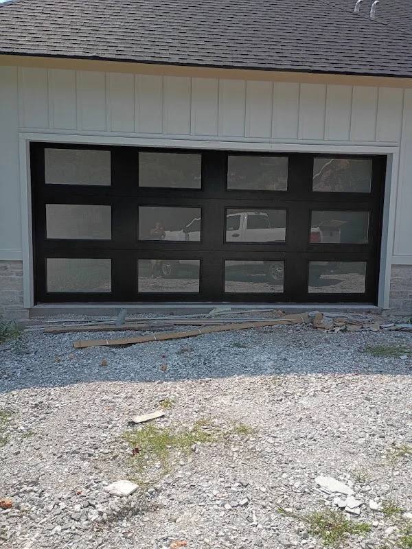 Elite Garage Door Services of Northwest ArkansasA newly installed black residential garage door with windows in a white house, surrounded by gravel and construction debris, showcasing professional garage door installation services in Northwest Arkansas.