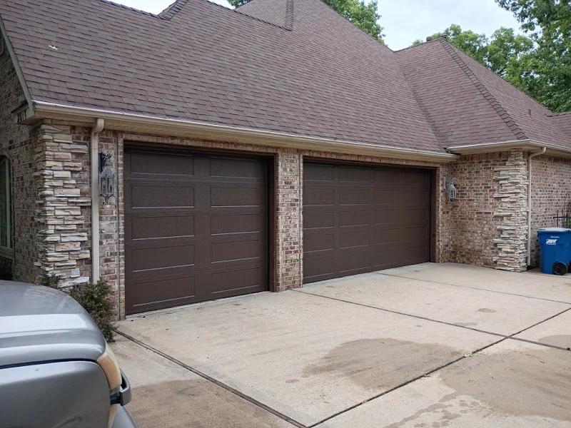 Elite Garage Door Services of Northwest ArkansasA brick house in Northwest Arkansas features two closed brown residential garage doors. A trash bin and part of a parked vehicle are visible. The image highlights the need for professional garage door service in NWA.