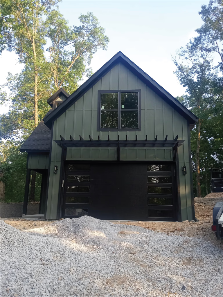 Elite Garage Door Services of Northwest ArkansasA modern black residential garage door is installed on a green house surrounded by trees, exemplifying professional garage door service in Northwest Arkansas, with gravel piled in the foreground indicating recent construction work.