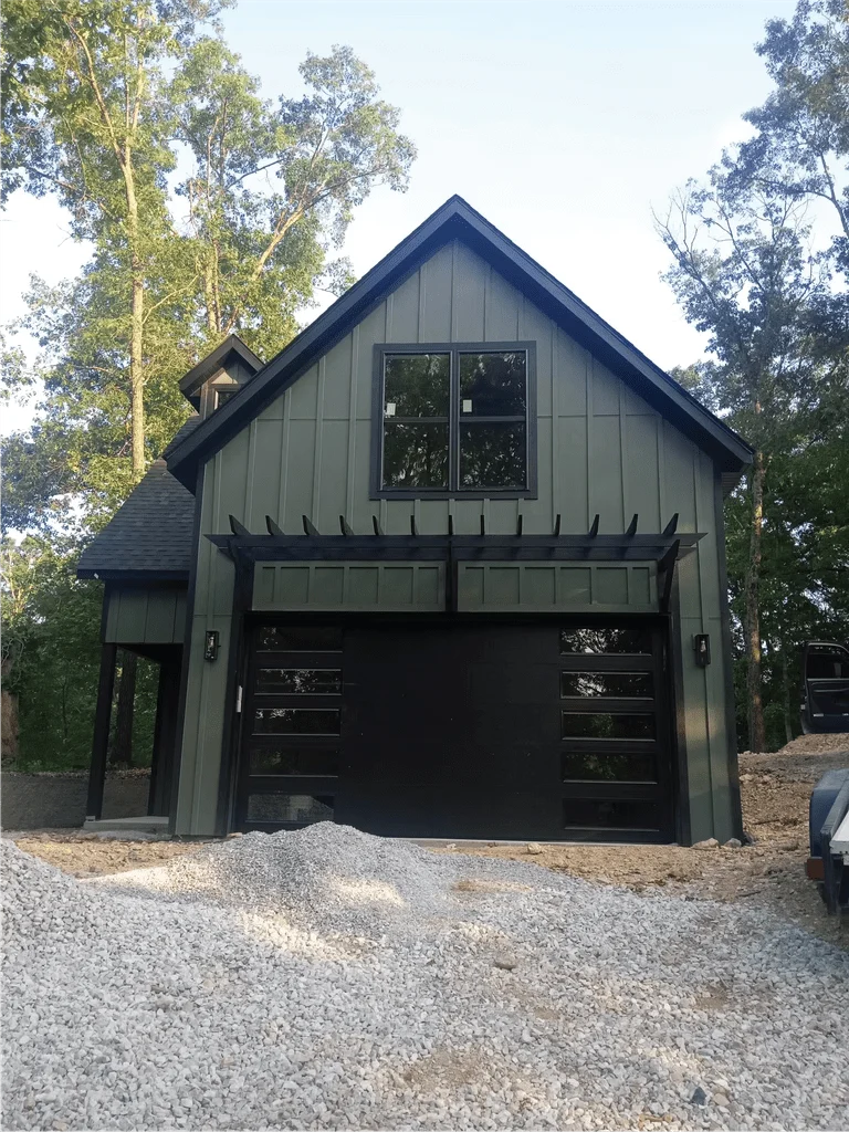Elite Garage Door Services of Northwest ArkansasA modern black residential garage door is installed on a green house surrounded by trees, exemplifying professional garage door service in Northwest Arkansas, with gravel piled in the foreground indicating recent construction work.