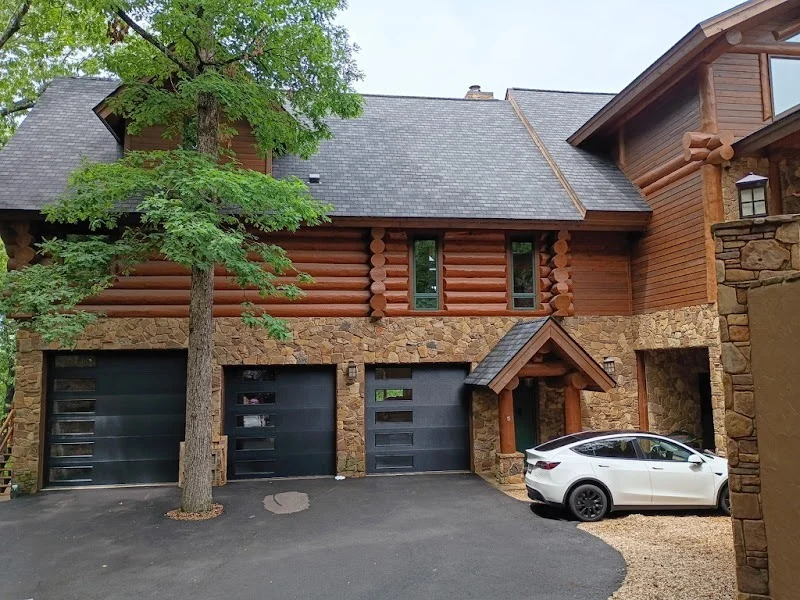 Elite Garage Door Services of Northwest ArkansasA rustic log cabin with three modern black overhead garage doors, surrounded by trees. A white car is parked nearby. This scene reflects the charm of residential areas in Northwest Arkansas, ideal for reliable garage door service.