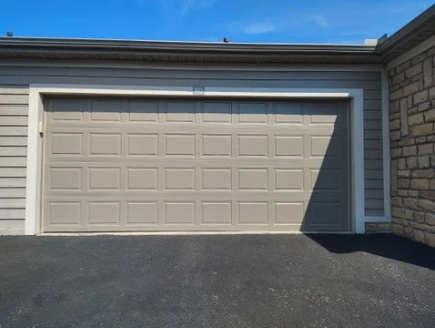 Elite Garage Door Services of Northwest ArkansasA tan residential garage door is closed, casting a shadow on the black asphalt driveway. The image depicts a typical home in Northwest Arkansas, highlighting the reliability of local garage door service providers.