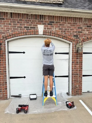 Elite Garage Door Services of Northwest ArkansasA professional technician from Elite Garage Door stands on a ladder, performing garage door repair on a white residential garage in Rogers, AR. Tools are scattered on the ground, indicating a reliable local service in Northwest Arkansas.