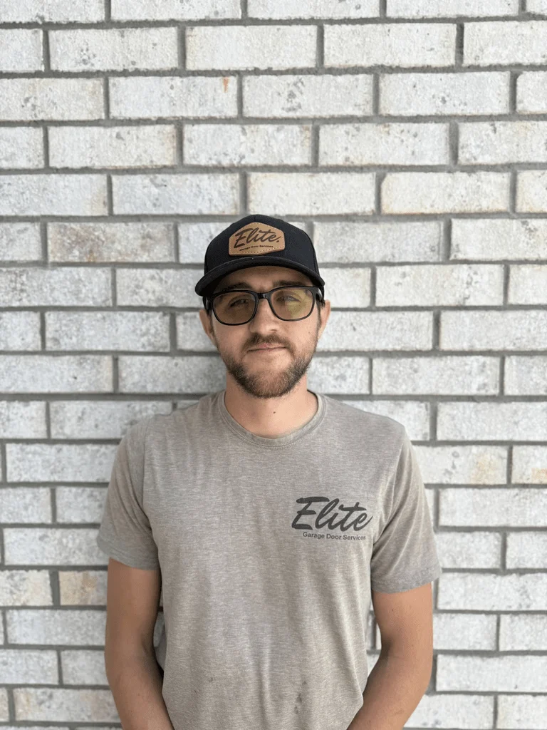 Elite Garage Door Services of Northwest ArkansasA professional technician from Elite Garage Door stands against a light brick wall, wearing a cap and shirt with the company logo, representing reliable garage door service in Northwest Arkansas.