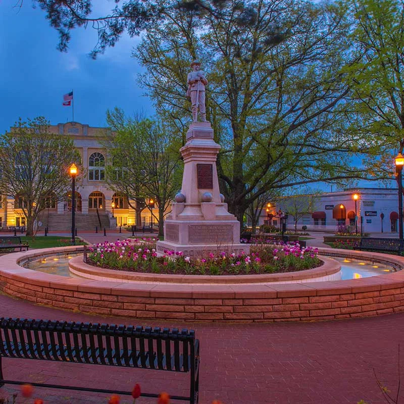 Elite Garage Door Services of Northwest Arkansas-A scenic view of a historic monument surrounded by blooming flowers and trees in a town square at dusk, with soft lighting from street lamps. This picturesque setting is in Bentonville AR, highlighting the charm of Northwest Arkansas.