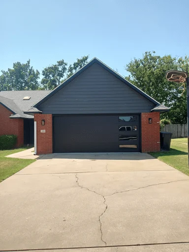 Elite Garage Door Services of Northwest ArkansasA modern residential garage in Northwest Arkansas features a sleek, dark overhead garage door, emphasizing local garage door installation and repair services. The house is brick with a concrete driveway and a basketball hoop on the side.