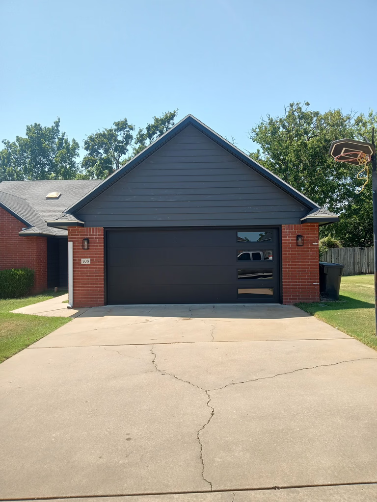 Elite Garage Door Services of Northwest ArkansasA modern black residential garage door is featured on a brick house in a suburban neighborhood, under a clear blue sky. The image represents reliable garage door service available in Northwest Arkansas.