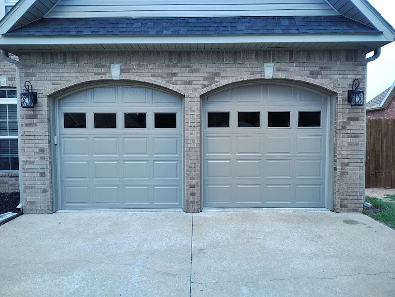 Elite Garage Door Services of Northwest ArkansasTwo beige residential garage doors in a brick house, showcasing a recent garage door installation in Northwest Arkansas. The driveway is clean, emphasizing professional and reliable service.