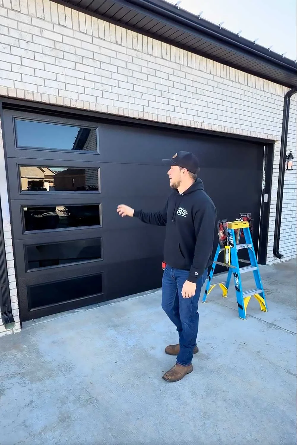 Elite Garage Door Services of Northwest Arkansas-A professional technician in a black hoodie stands beside a modern black residential garage door, discussing garage door installation in Northwest Arkansas. A blue ladder and tools are visible, highlighting reliable garage door service.