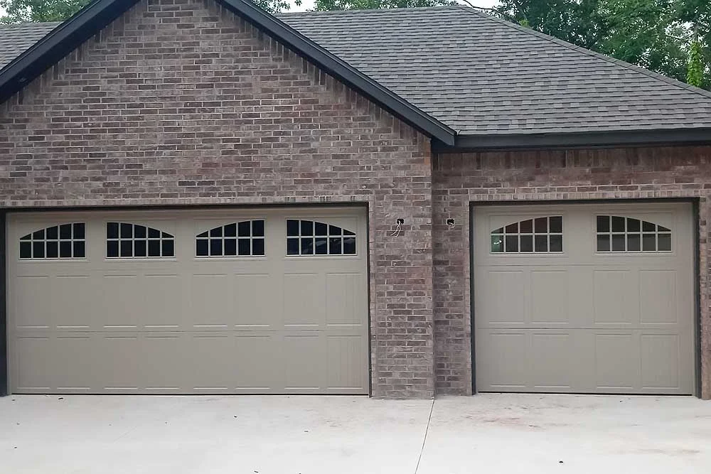 Elite Garage Door Services of Northwest Arkansas-A brick house in Northwest Arkansas features two beige residential garage doors with decorative windows, highlighting a recent garage door installation showcasing local craftsmanship and reliable service.
