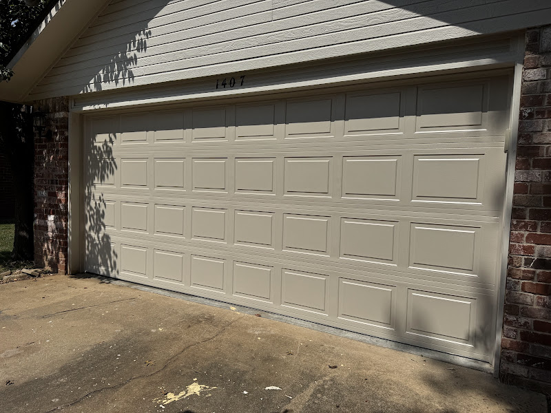 Elite Garage Door Services of Northwest ArkansasA beige residential garage door, recently installed, is closed in a driveway in Northwest Arkansas, highlighting local professional garage door installation services.