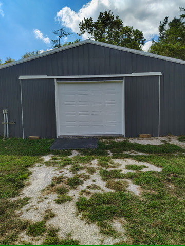 Elite Garage Door Services of Northwest ArkansasA white residential garage door is installed in a gray building surrounded by greenery under a partly cloudy sky, illustrating professional garage door installation services available in Northwest Arkansas.