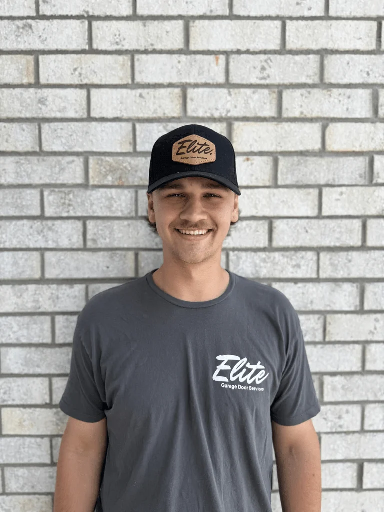 Elite Garage Door Services of Northwest ArkansasA smiling technician from Elite Garage Door Services, wearing a branded shirt and cap, stands against a brick wall, representing professional and reliable garage door service in Northwest Arkansas.