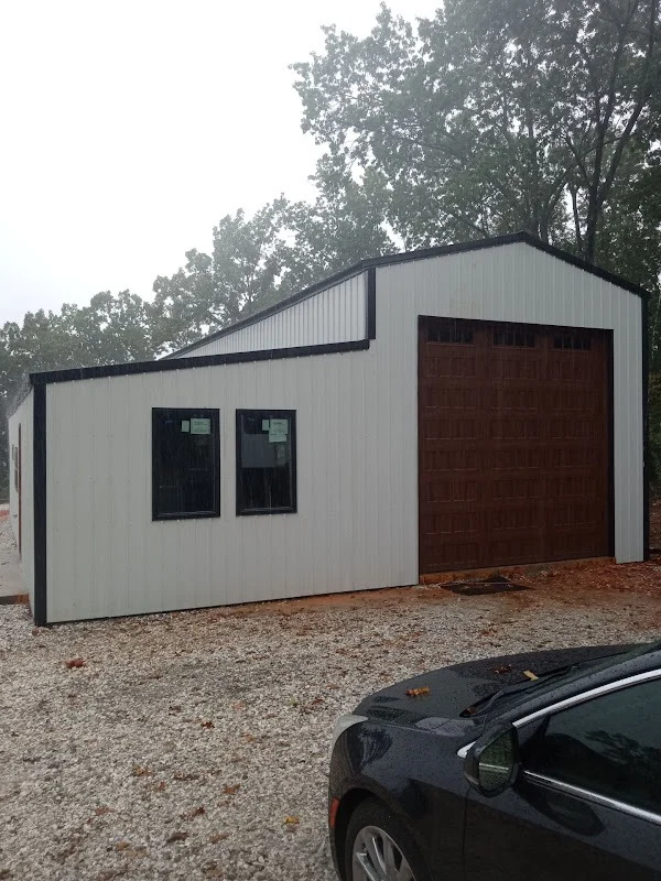 Elite Garage Door Services of Northwest ArkansasA modern building with a large brown overhead garage door, surrounded by trees in a gravel area, is seen on a rainy day in Northwest Arkansas. A parked car is partially visible in the foreground.