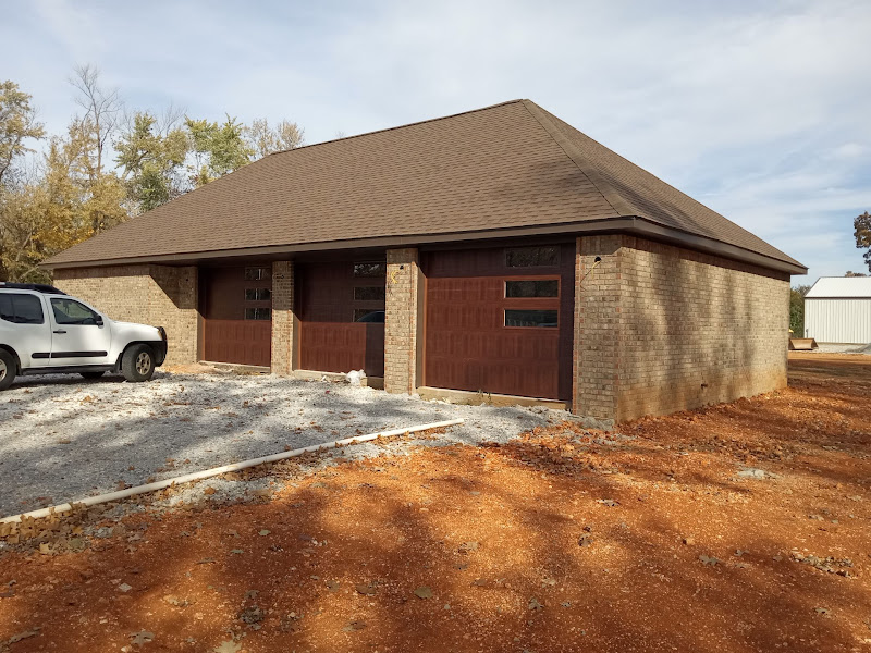 Elite Garage Door Services of Northwest ArkansasA brick residential garage with three brown overhead doors and a white truck parked beside it, located in Northwest Arkansas. The setting suggests professional garage door installation services in a new construction area.