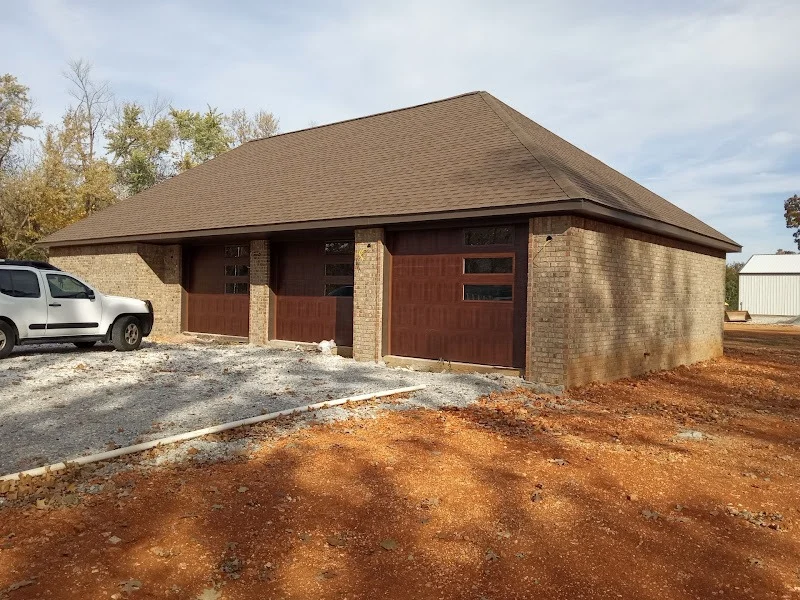 Elite Garage Door Services of Northwest ArkansasA brick residential garage with three brown overhead doors and a white truck parked beside it, located in Northwest Arkansas. The setting suggests professional garage door installation services in a new construction area.