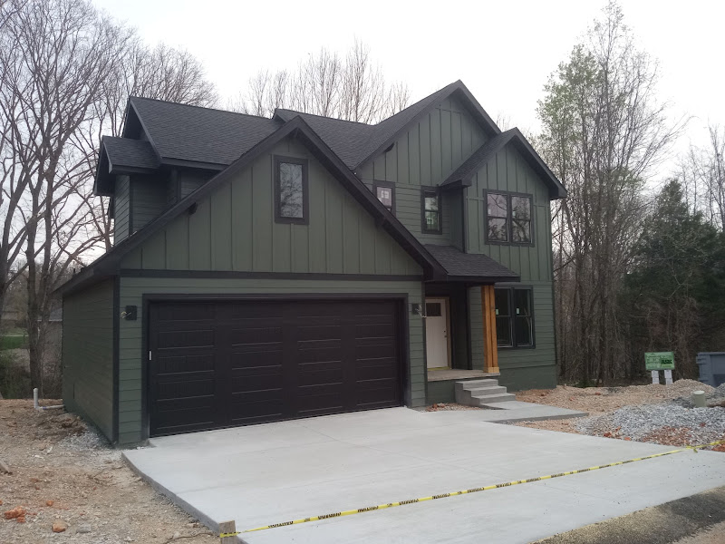 Elite Garage Door Services of Northwest ArkansasA newly constructed dark green house in Northwest Arkansas with a double-car garage featuring sleek, black residential garage doors, surrounded by trees and a gravel yard, ideal for professional garage door service in the NWA region.