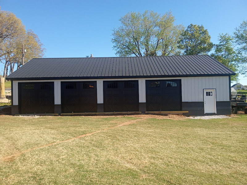 Elite Garage Door Services of Northwest ArkansasA large, modern garage with multiple black overhead garage doors is set against a backdrop of trees on a sunny day. The building is part of a residential setup in Northwest Arkansas, showcasing a recent garage door installation.