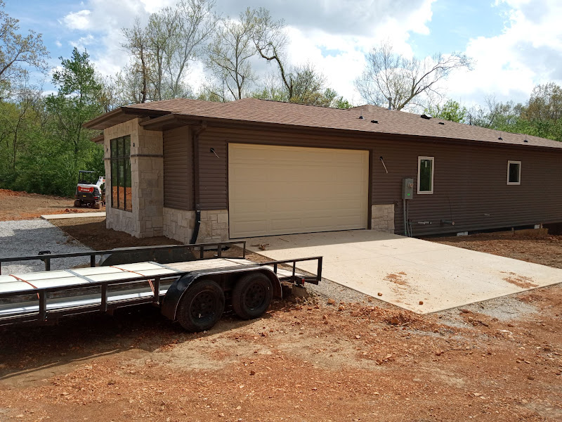 Elite Garage Door Services of Northwest ArkansasA newly constructed home in Northwest Arkansas features a beige residential garage door with a nearby trailer. The setting is surrounded by trees under a…