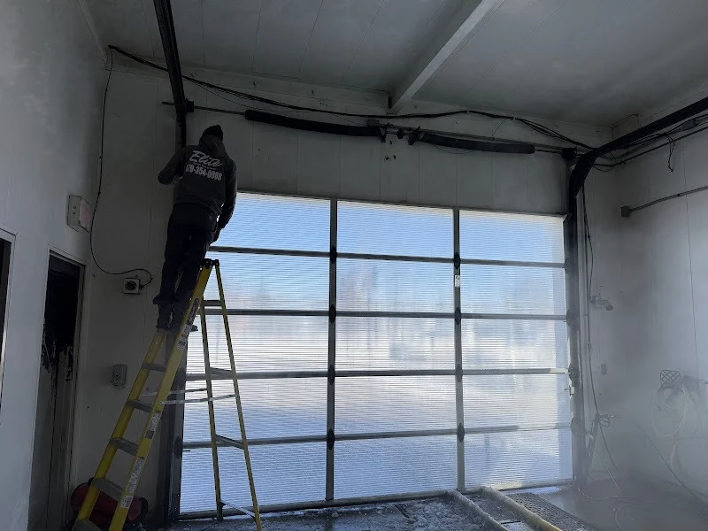 Elite Garage Door Services of Northwest ArkansasA professional technician stands on a ladder, repairing garage door springs inside a garage with overhead doors in Northwest Arkansas. Sunlight filters through the translucent panels, highlighting the local, reliable garage door service in progress.