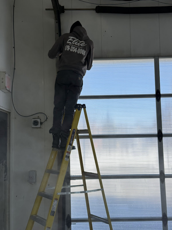 Elite Garage Door Services of Northwest ArkansasA professional technician from Elite Garage Door is on a ladder inspecting garage door springs inside a building with overhead garage doors, showcasing reliable garage door service in Northwest Arkansas.