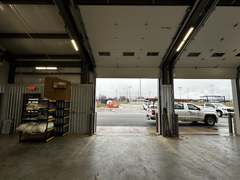 Elite Garage Door Services of Northwest ArkansasInside a garage with open doors, Elite Garage Door's branded trucks are parked outside, ready for garage door service in Northwest Arkansas. Shelves with supplies are visible on the left, indicating a professional and organized workspace.