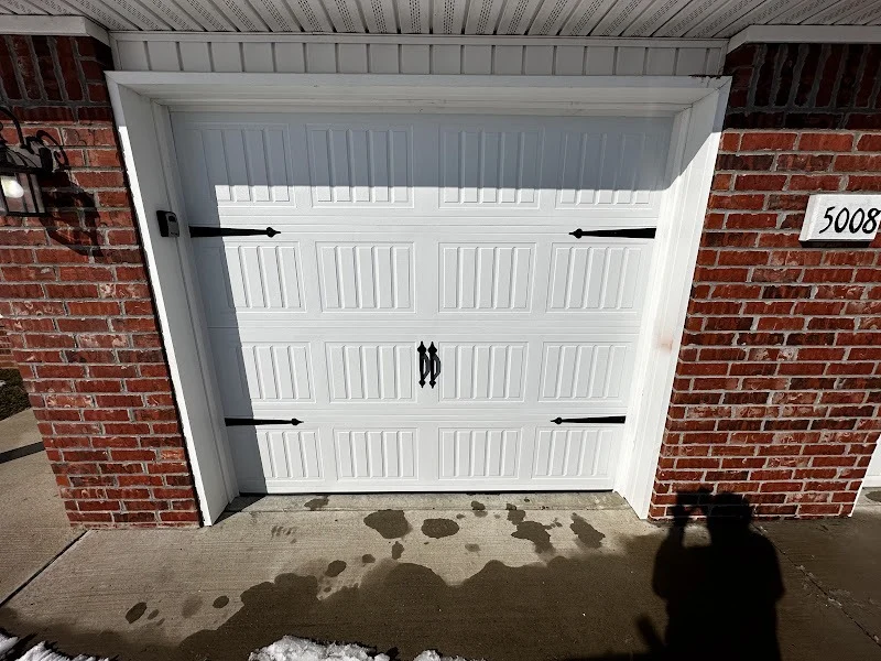 Elite Garage Door Services of Northwest ArkansasA white residential garage door with decorative black handles and hinges is set within a red brick exterior. The scene suggests a recent garage door…
