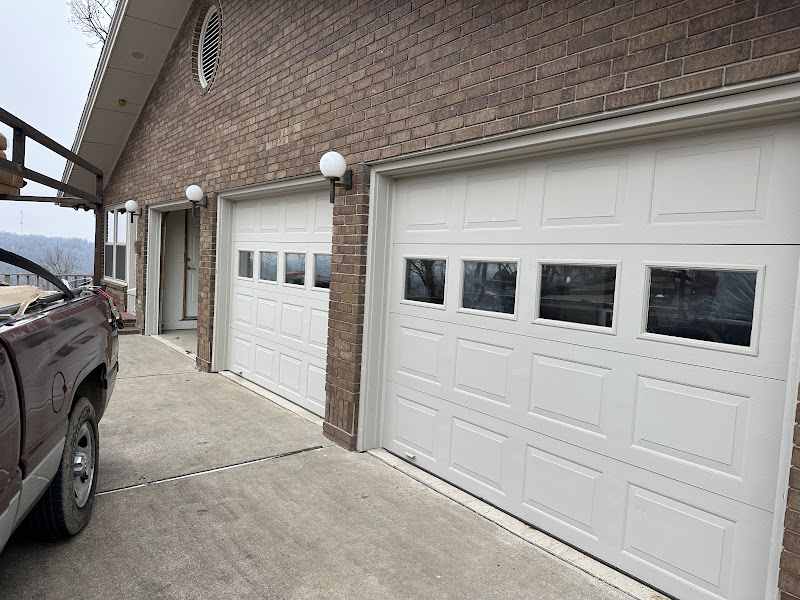 Elite Garage Door Services of Northwest ArkansasWhite residential garage doors with windows are installed on a brick home, with a local service truck parked nearby, highlighting reliable garage door service in Northwest Arkansas.