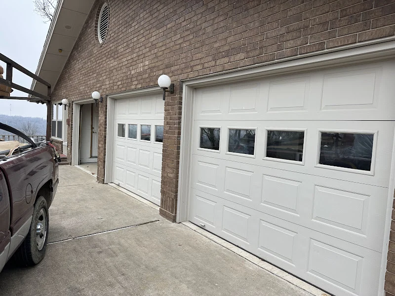 Elite Garage Door Services of Northwest ArkansasWhite residential garage doors with windows are installed on a brick home, with a local service truck parked nearby, highlighting reliable garage door service in Northwest Arkansas.