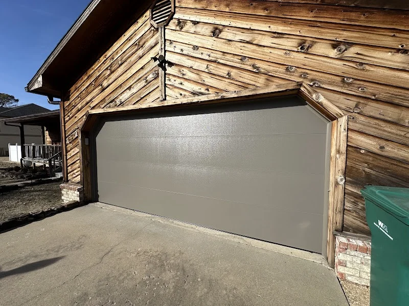 Elite Garage Door Services of Northwest ArkansasA newly installed, gray residential garage door is shown on a wooden house in Rogers AR, highlighting the quality of local garage door service in Northwest Arkansas.