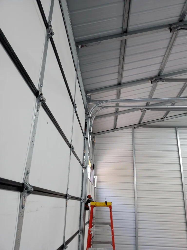 Elite Garage Door Services of Northwest ArkansasA professional technician stands on a red ladder performing garage door repair on overhead garage doors in a spacious, well-lit interior in Northwest Arkansas, emphasizing local, reliable service.