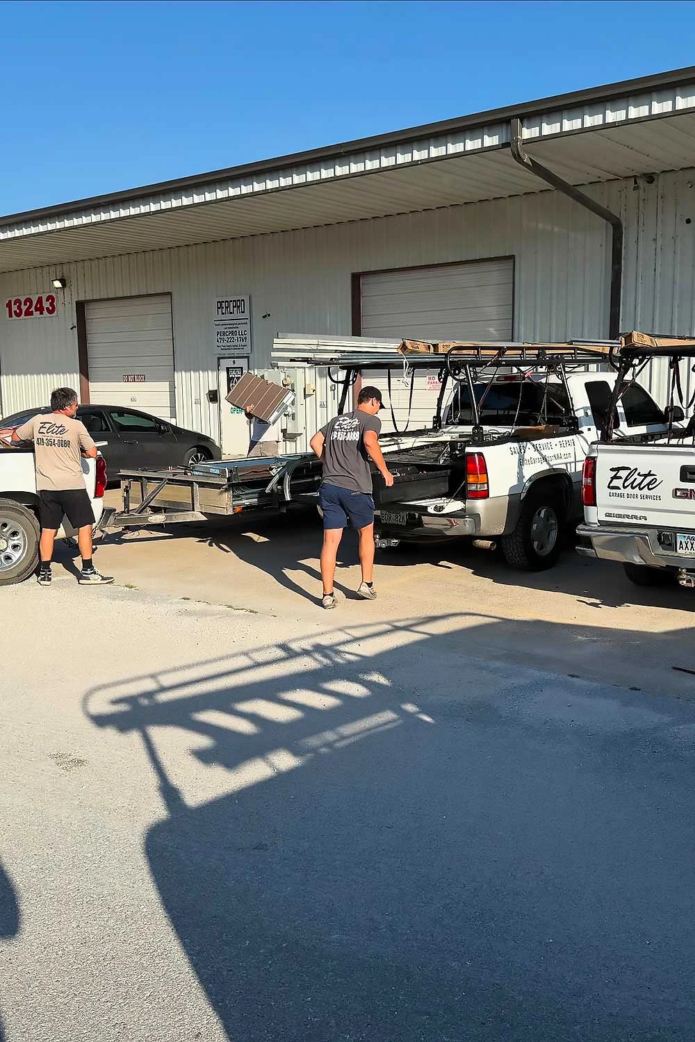 Elite Garage Door Services of Northwest Arkansas-Two technicians from Elite Garage Door load equipment onto trucks outside a warehouse in Rogers AR, preparing for garage door service. The vehicles display the…