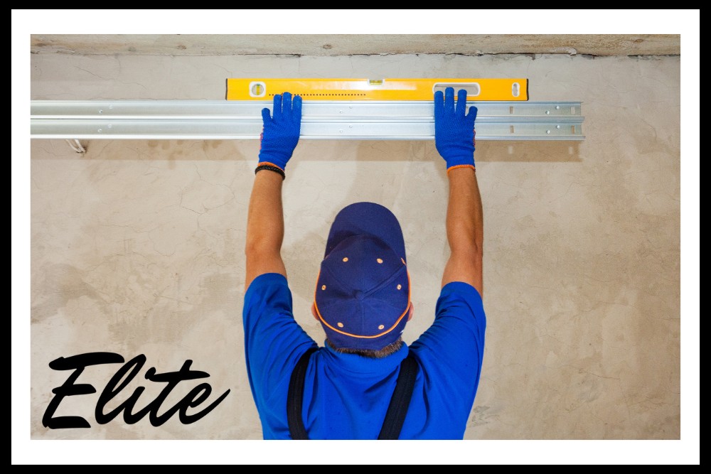 Why Regular Garage Door Tune-Ups Save You Money-1 Elite Garage Door Services of Northwest Arkansas-A professional technician in blue uniform and gloves installs an overhead garage door track using a level, illustrating reliable garage door installation services in Northwest Arkansas.