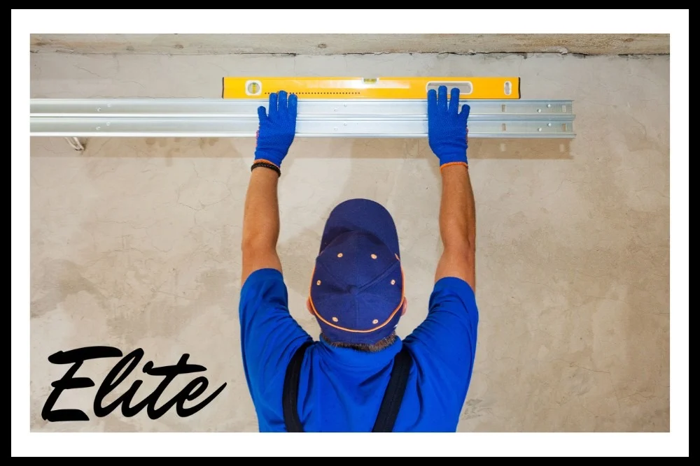Why Regular Garage Door Tune-Ups Save You Money-1 Elite Garage Door Services of Northwest Arkansas-A professional technician in blue uniform and gloves installs an overhead garage door track using a level, illustrating reliable garage door installation services in Northwest Arkansas.