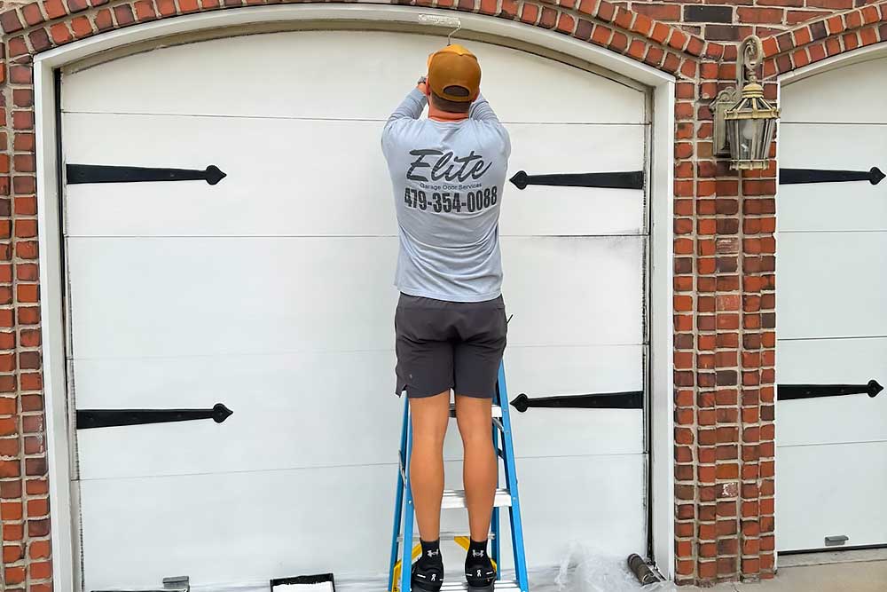 Elite Garage Door Services of Northwest Arkansas-A professional technician from Elite Garage Door is on a ladder performing garage door repair on a white overhead garage door in Northwest Arkansas, showcasing reliable local service.