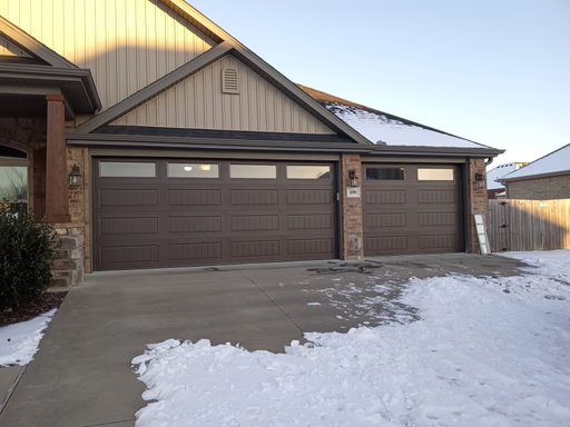 Elite Garage Door Services of Northwest ArkansasA snow-covered driveway leads to a home with two large, brown residential garage doors in Northwest Arkansas, suggesting reliable garage door service. The winter setting highlights the need for professional maintenance.