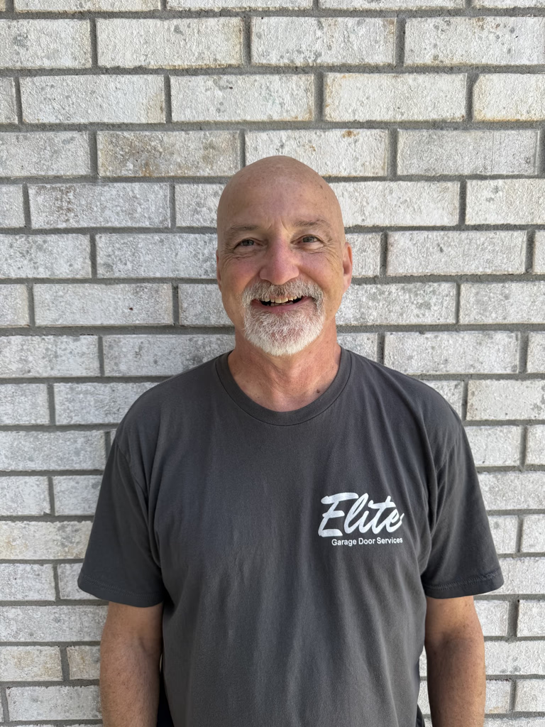 Elite Garage Door Services of Northwest ArkansasA smiling man in a gray "Elite Garage Door Services" shirt stands against a light brick wall, representing professional and reliable garage door repair in Northwest Arkansas.